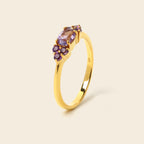 A yellow gold ring featuring a central oval purple gemstone surrounded by small round purple accent stones, set on a smooth gold band against a light beige background.