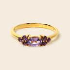 A yellow gold ring featuring a central oval-cut purple amethyst gemstone flanked by two clusters of smaller round purple amethyst stones, set against a solid light beige background.