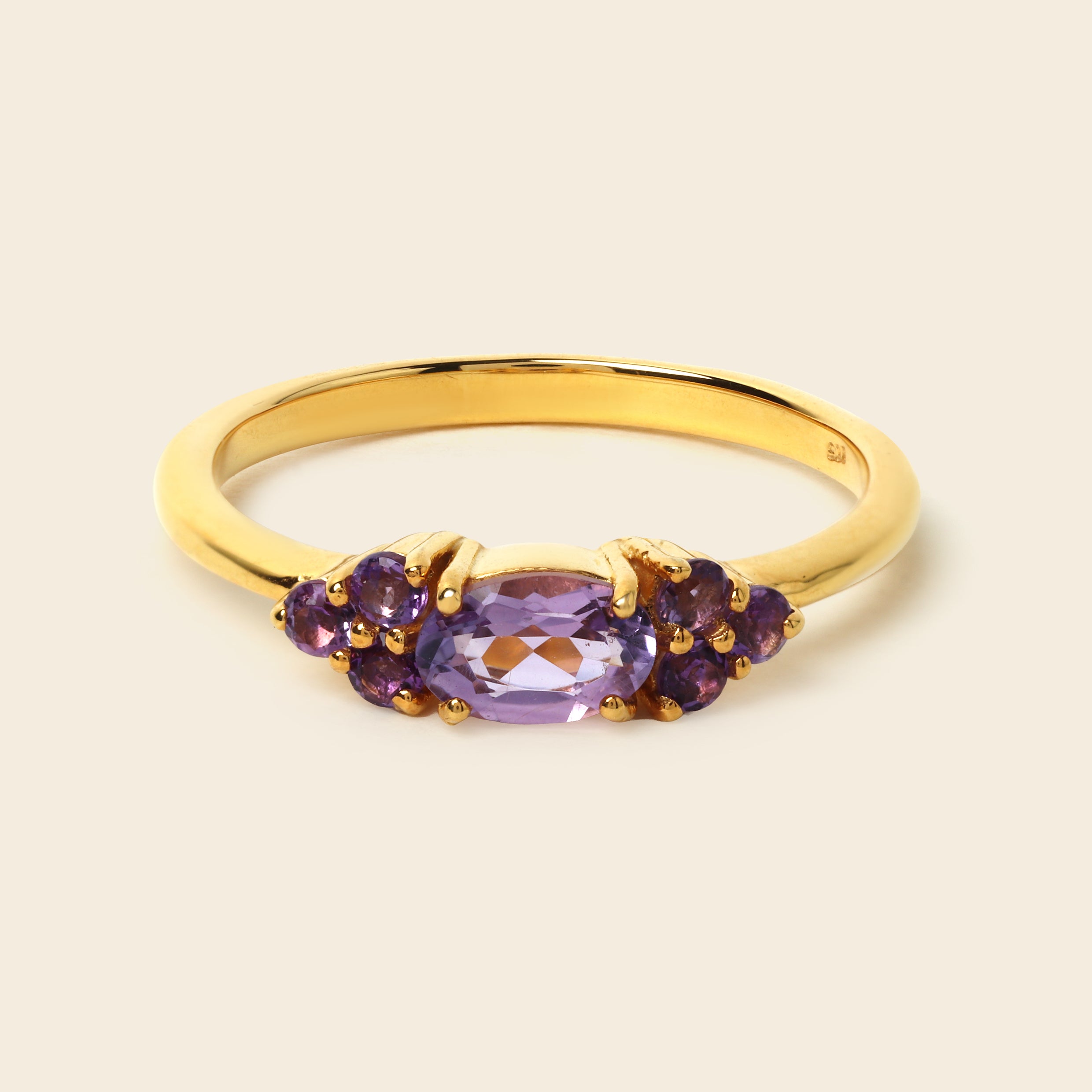 A yellow gold ring featuring a central oval-cut purple amethyst gemstone flanked by two clusters of smaller round purple amethyst stones, set against a solid light beige background.