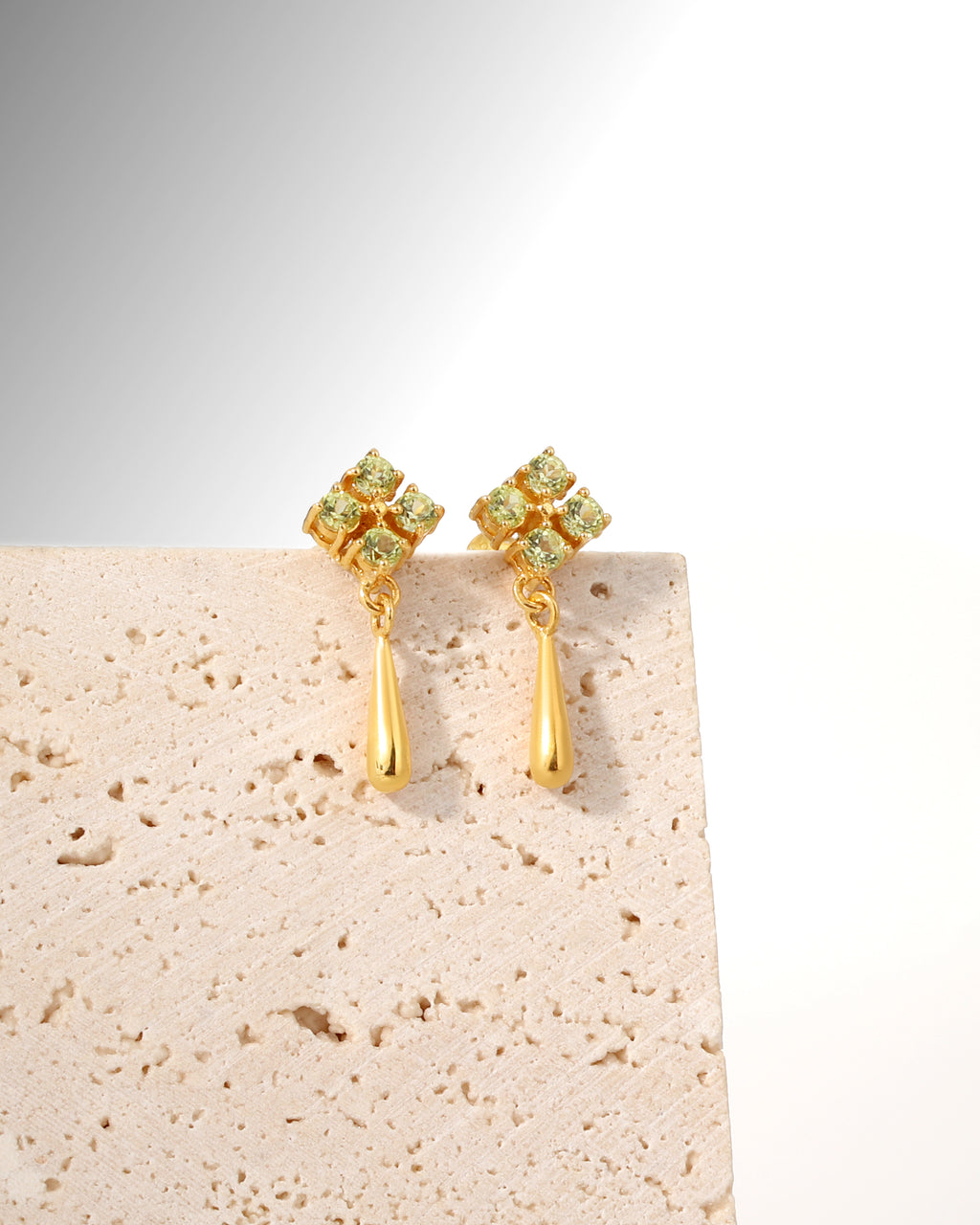 A pair of gold dangle earrings with a small cluster of pale green gemstones at the top and a teardrop-shaped gold drop below, displayed on a textured beige stone block against a white background.