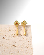 A pair of gold dangle earrings with a small cluster of pale green gemstones at the top and a teardrop-shaped gold drop below, displayed on a textured beige stone block against a white background.