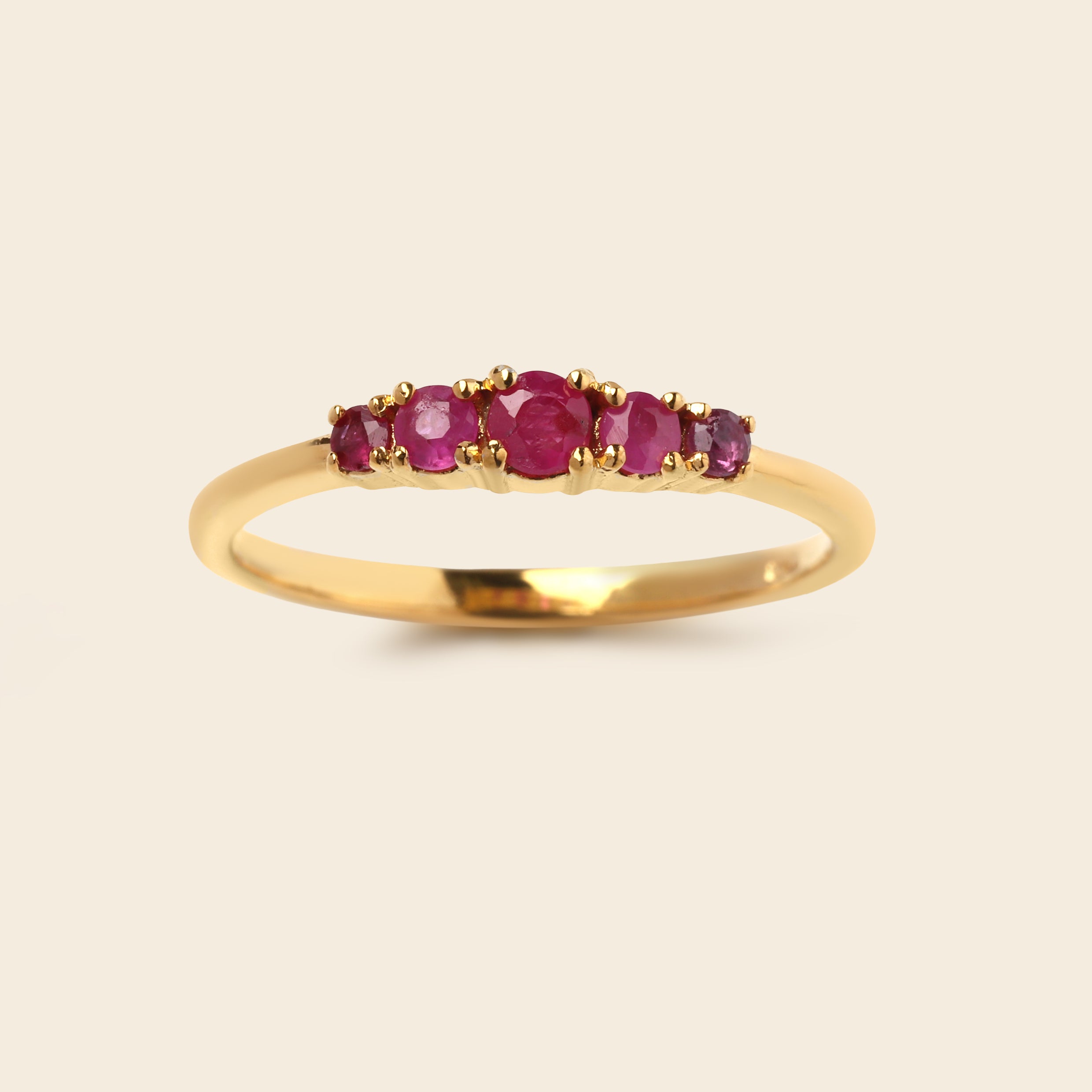 A yellow gold ring featuring five round-cut pinkish-red gemstones set across the top, presented on a light beige background.