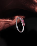 A silver band ring featuring a row of small round pink gemstones, displayed against a dark background with a draped copper-brown fabric accent.