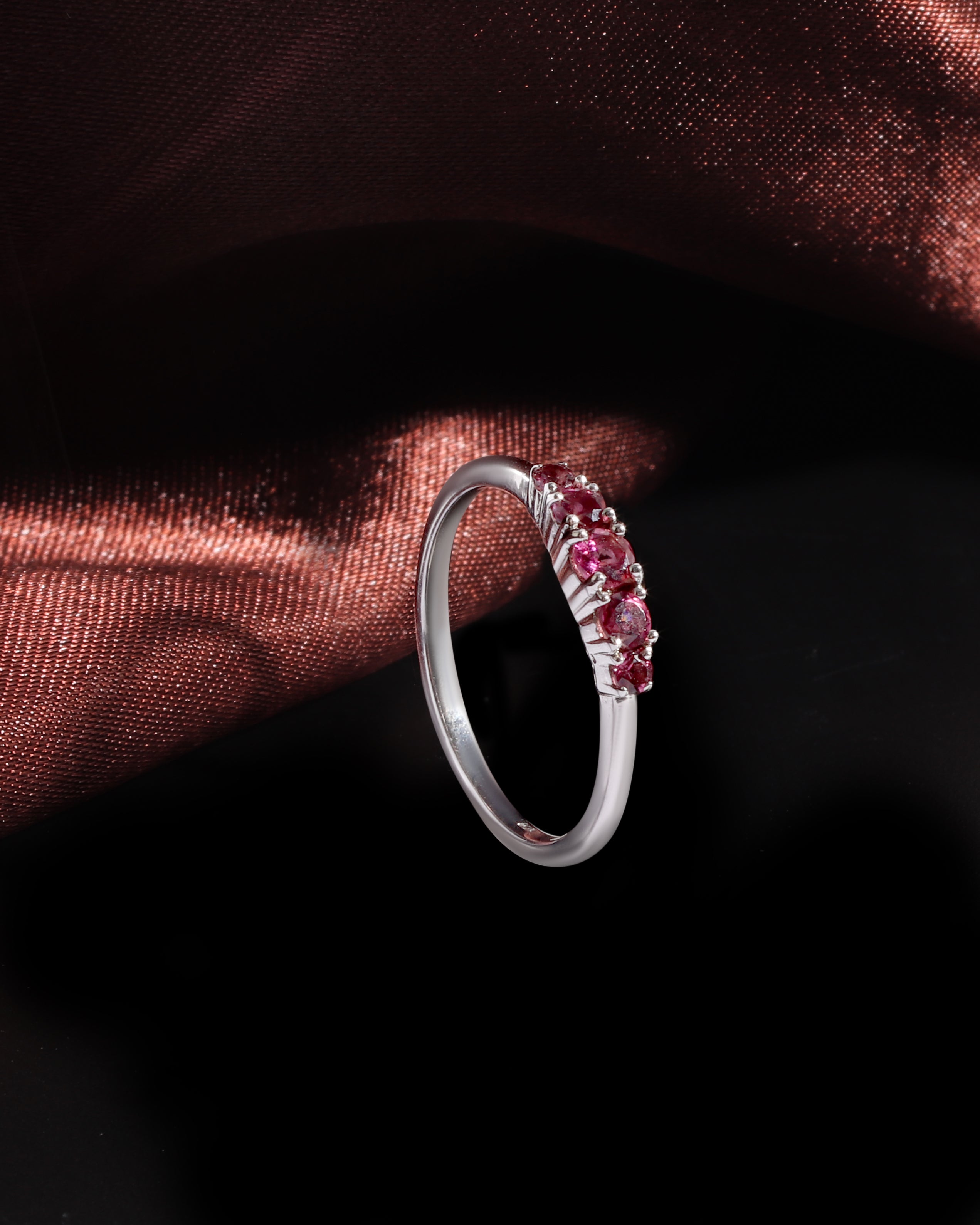 A silver band ring featuring a row of small round pink gemstones, displayed against a dark background with a draped copper-brown fabric accent.