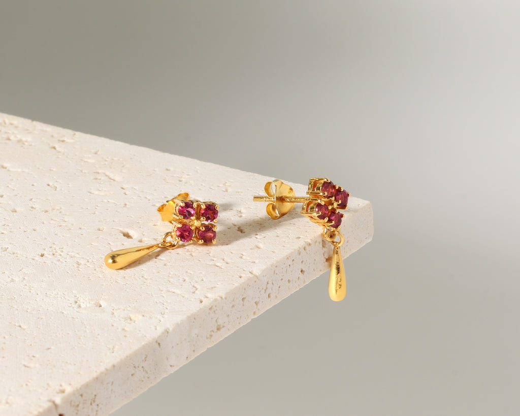 A pair of gold dangle earrings with small clusters of round red gemstones and teardrop-shaped gold drops, displayed on the corner of a light stone block against a soft gray background.