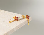 A pair of gold dangle earrings with small clusters of round red gemstones and teardrop-shaped gold drops, displayed on the corner of a light stone block against a soft gray background.