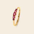 A gold band ring featuring a row of five oval-cut pinkish-red gemstones, set against a solid light beige background.