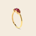 A gold ring featuring a large oval-cut pinkish-red center stone and two smaller round stones on the side, shown on a light beige background.
