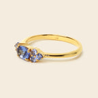 A yellow gold ring featuring a central oval blue gemstone flanked by two smaller pear-cut blue gemstones, set against a solid light beige background.
