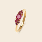 A yellow gold ring featuring a cluster of oval and round deep pinkish-red gemstones, set against a solid light beige background.