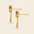 A pair of gold drop earrings with cylindrical studs featuring small blue inlay details, shown on a light beige background.