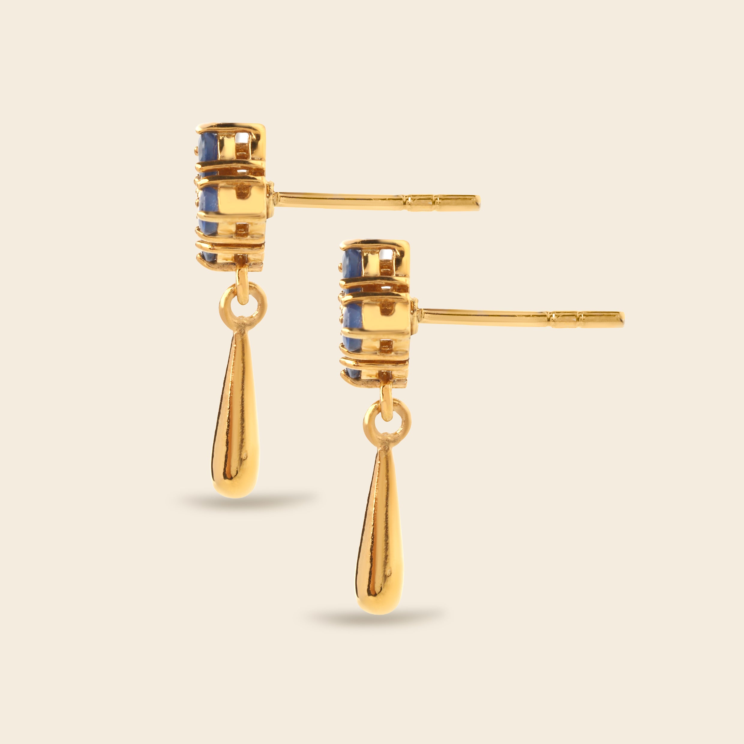 A pair of gold drop earrings with cylindrical studs featuring small blue inlay details, shown on a light beige background.