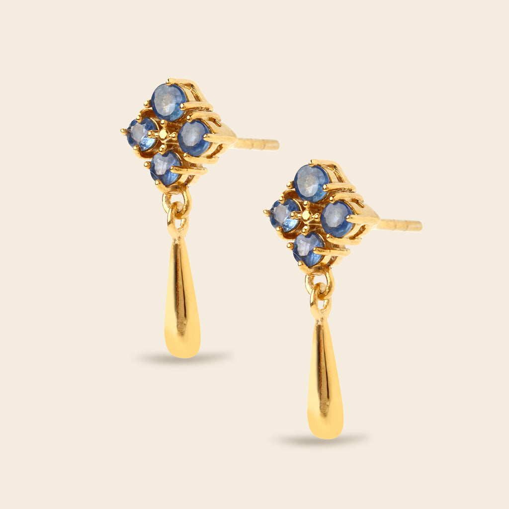 A pair of gold dangle earrings with a floral cluster of round blue gemstones and a teardrop-shaped gold drop, shown on a light beige background.