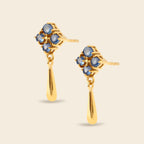 A pair of gold dangle earrings with a floral cluster of round blue gemstones and a teardrop-shaped gold drop, shown on a light beige background.