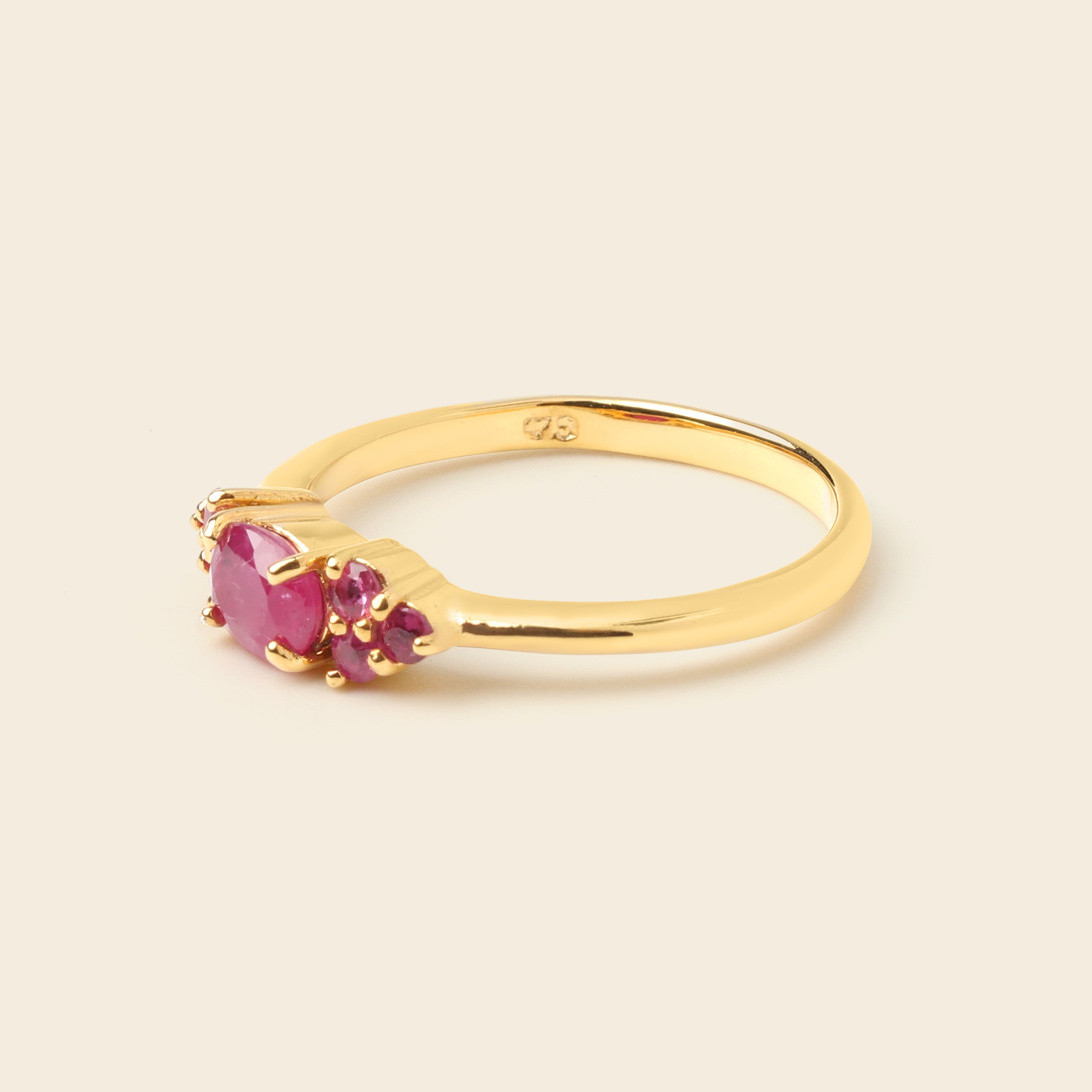 A yellow gold ring featuring a large oval pink gemstone and two smaller round-cut pink gemstones, set in a polished gold band against a light beige background.