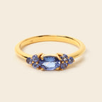A yellow gold ring featuring a central oval-cut blue gemstone flanked by smaller round blue gemstones, set against a solid light beige background.