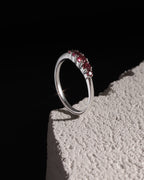 A silver ring with a row of small red gemstones, standing on a textured white block against a solid black background.