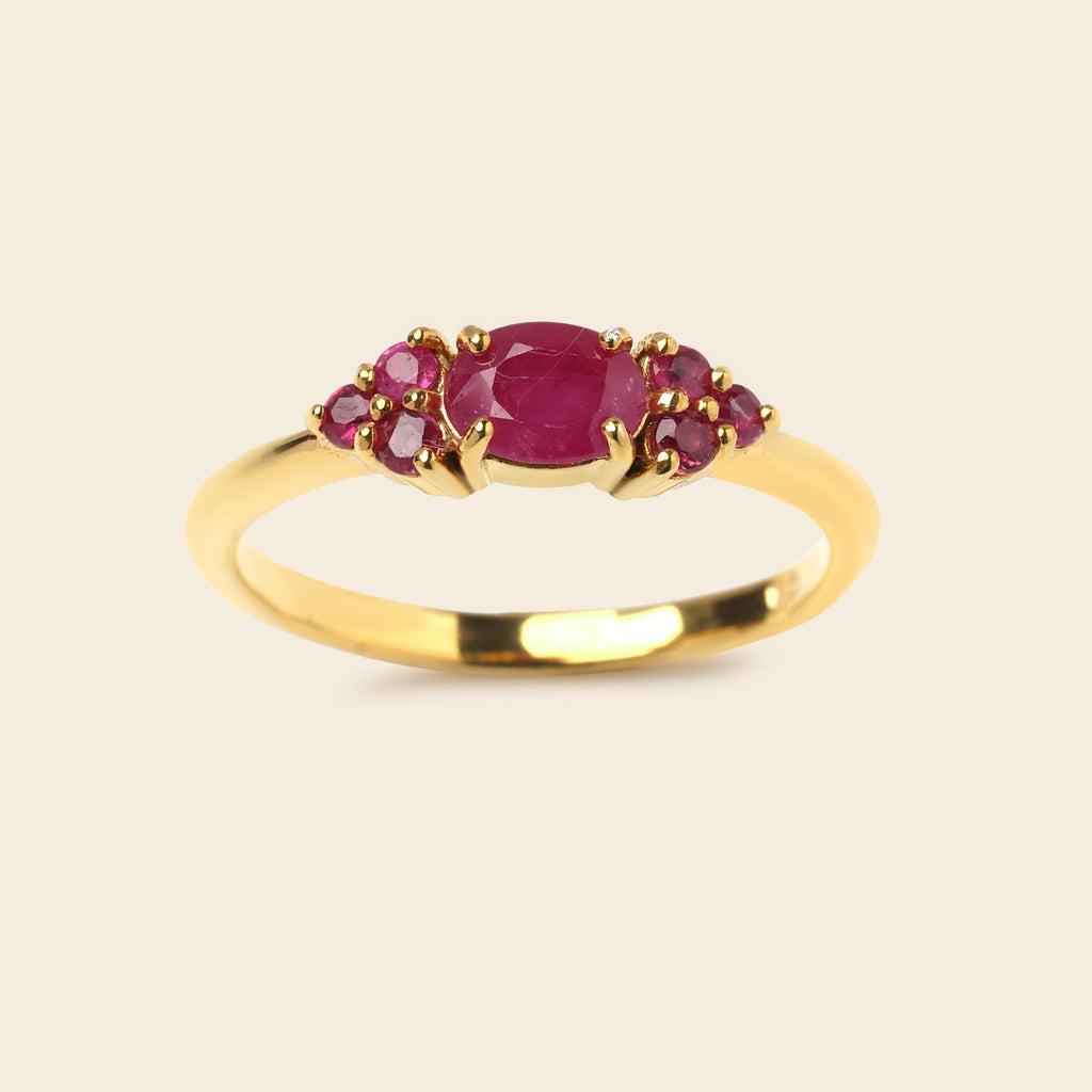 A yellow gold ring featuring a central oval pinkish-red ruby gemstone flanked by two small clusters of round pinkish-red ruby gemstones, set against a solid light beige background.