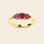 A yellow gold ring featuring a central oval pinkish-red ruby gemstone flanked by two small clusters of round pinkish-red ruby gemstones, set against a solid light beige background.