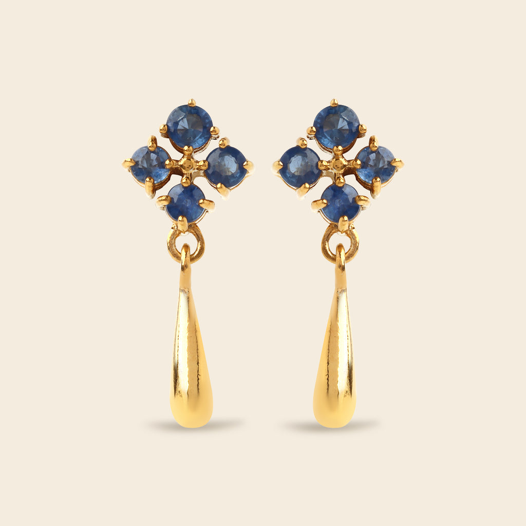 A pair of gold dangle earrings featuring a cluster of round blue gemstones at the top and a teardrop-shaped gold drop below, set against a solid light beige background.