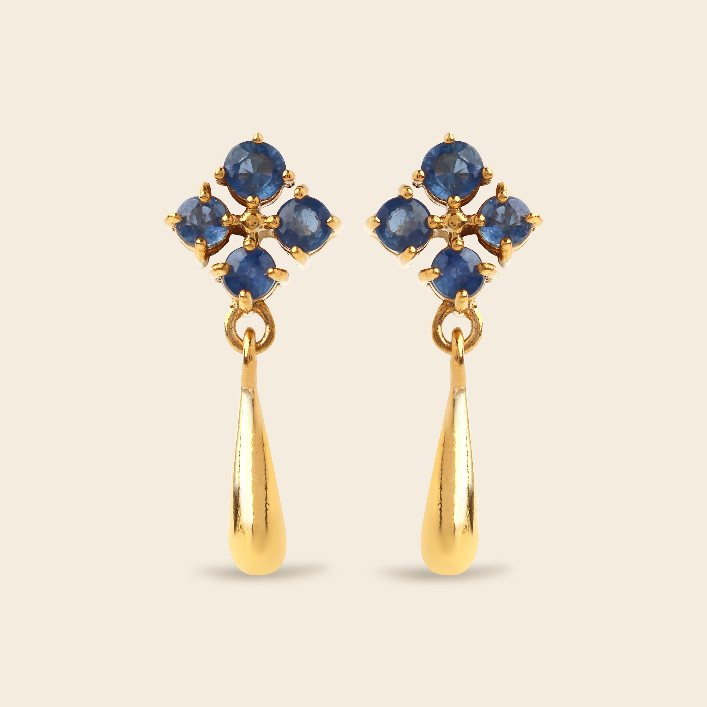 A pair of gold dangle earrings featuring a cluster of round blue gemstones at the top and a teardrop-shaped gold drop below, set against a solid light beige background.
