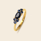 A gold ring featuring a central oval dark blue gemstone flanked by smaller round blue gemstones, set against a solid light beige background.