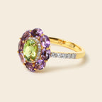 A yellow gold ring featuring a central oval green peridot gemstone surrounded by a cluster of round purple amethysts, with a pavé band set with small clear stones, shown on a light beige background.