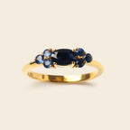 A gold ring featuring a central oval dark blue sapphire flanked by three smaller round blue sapphires, set against a solid light beige background.