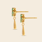 A pair of gold drop earrings with stacked rectangular gold settings featuring small green stones and teardrop-shaped gold dangles, shown on a light beige background.