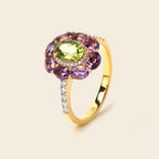 A yellow gold ring featuring a central oval green gemstone surrounded by a halo of purple oval gemstones and small clear accent stones, set against a light beige background.