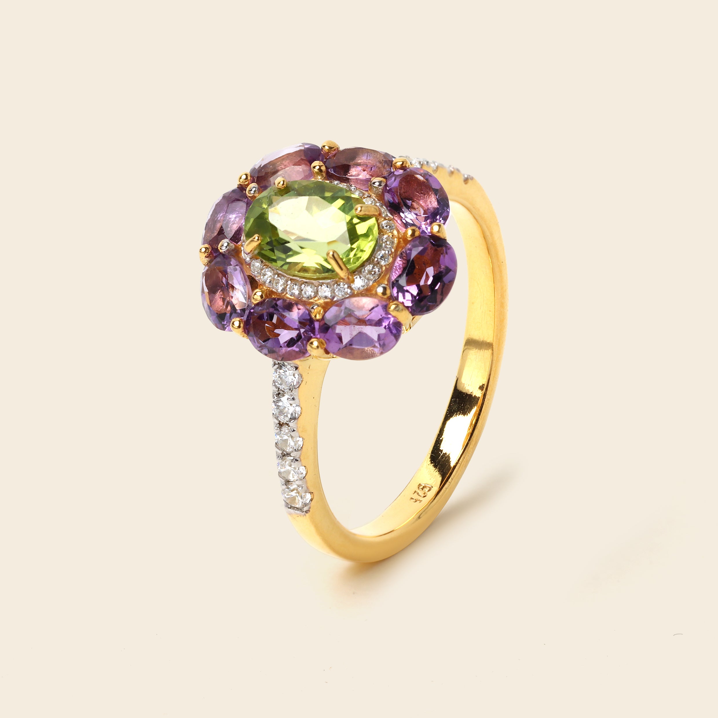 A yellow gold ring featuring a central oval green gemstone surrounded by a halo of purple oval gemstones and small clear accent stones, set against a light beige background.
