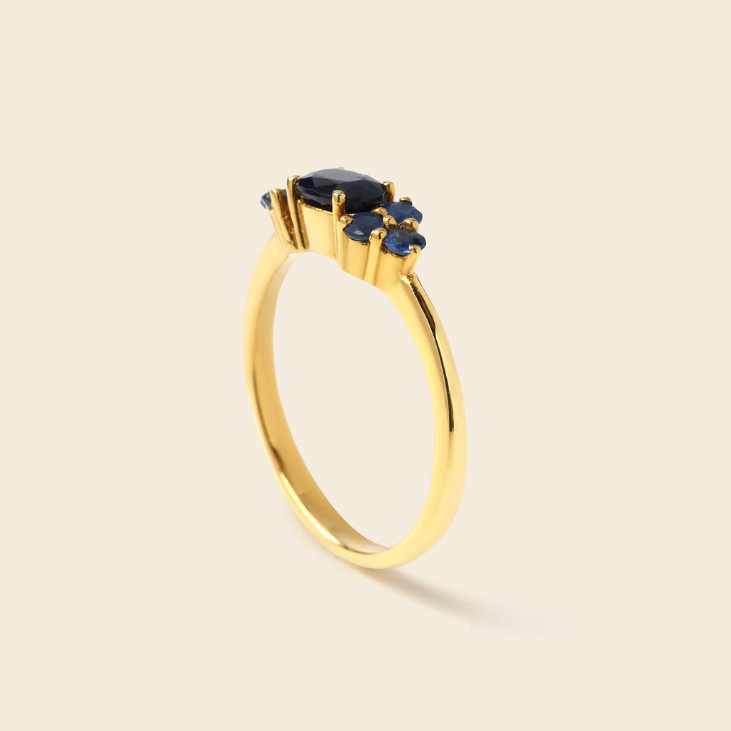 A gold ring featuring a central oval dark blue gemstone surrounded by three smaller round blue gemstones, set on a polished yellow gold band, shown on a light beige background.