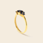 A gold ring featuring a central oval dark blue gemstone surrounded by three smaller round blue gemstones, set on a polished yellow gold band, shown on a light beige background.