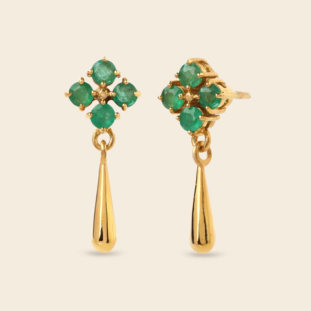 A pair of gold dangle earrings featuring a four-stone green gemstone floral cluster on the top and a polished gold teardrop drop below, set against a solid light beige background.