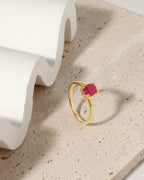 A gold ring with a round pink gemstone, displayed on a textured beige stone surface next to a white wavy ceramic prop.