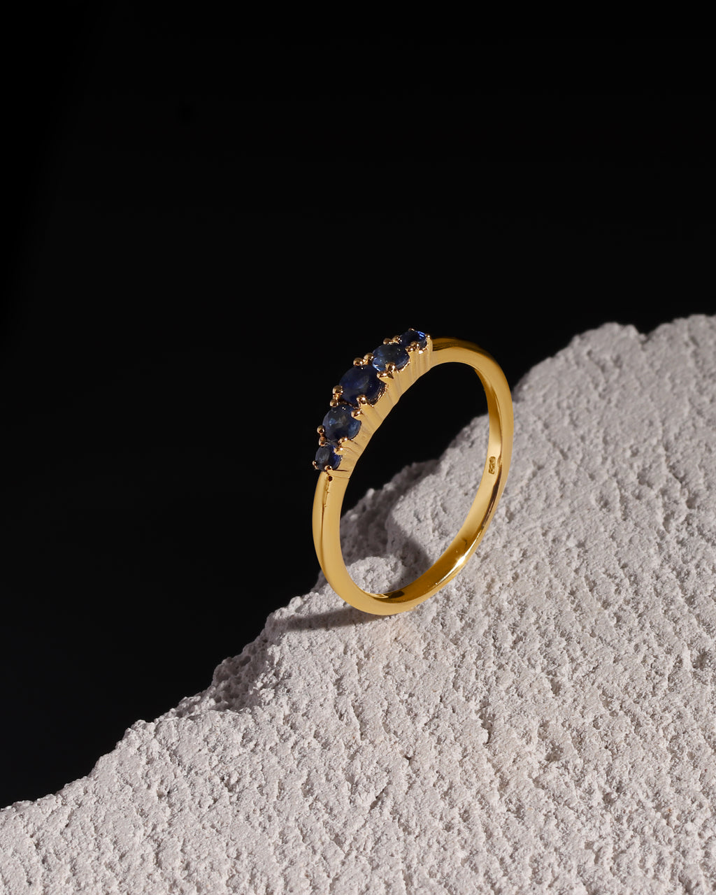 A gold band ring featuring a row of small dark blue gemstones, displayed on a textured white surface against a solid black background.