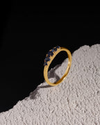 A gold band ring featuring a row of small dark blue gemstones, displayed on a textured white surface against a solid black background.