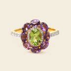 A yellow gold ring featuring a central oval green peridot gemstone surrounded by a halo of round purple amethyst stones, with a diamond-accented band, set against a solid light beige background.