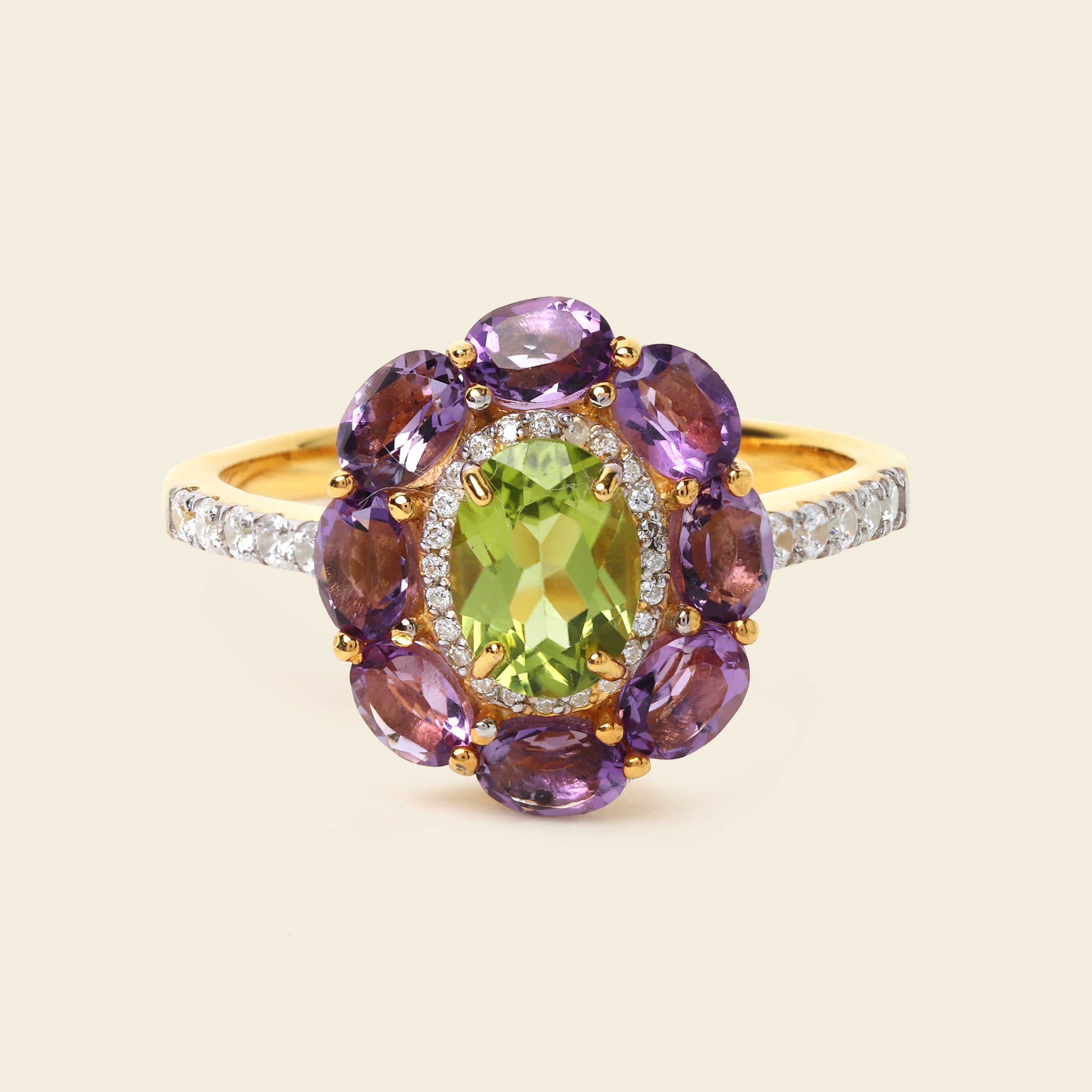 A yellow gold ring featuring a central oval green peridot gemstone surrounded by a halo of round purple amethyst stones, with a diamond-accented band, set against a solid light beige background.