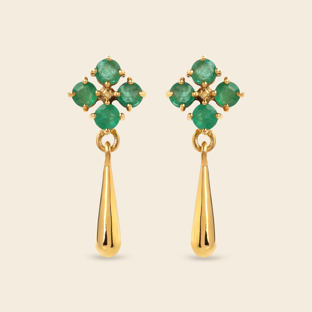 A pair of gold dangle earrings featuring a floral cluster of round green emerald-cut gemstones at the top and a polished gold teardrop drop below, set against a solid light beige background.