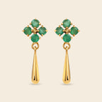 A pair of gold dangle earrings featuring a floral cluster of round green emerald-cut gemstones at the top and a polished gold teardrop drop below, set against a solid light beige background.