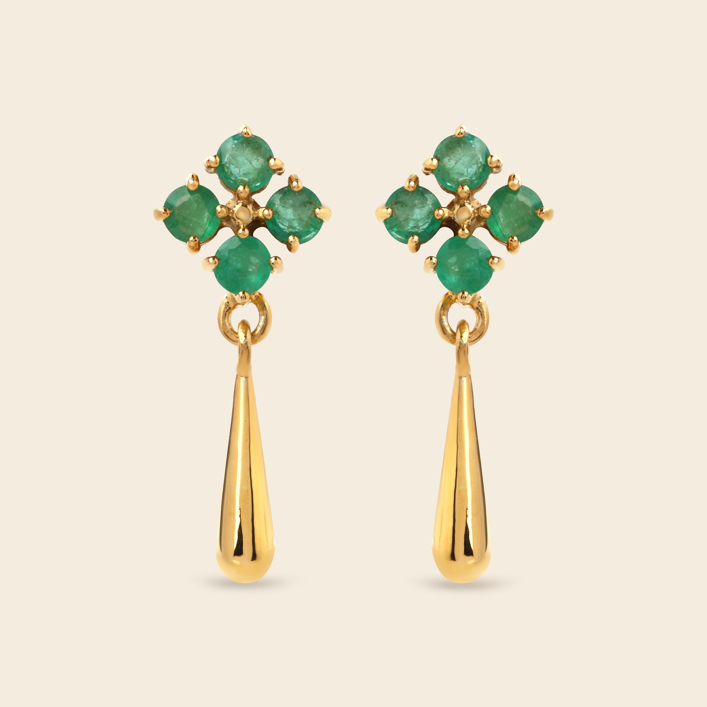 A pair of gold dangle earrings featuring a floral cluster of round green emerald-cut gemstones at the top and a polished gold teardrop drop below, set against a solid light beige background.