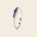 A silver ring featuring a row of round-cut blue gemstones, shown on a solid light beige background.