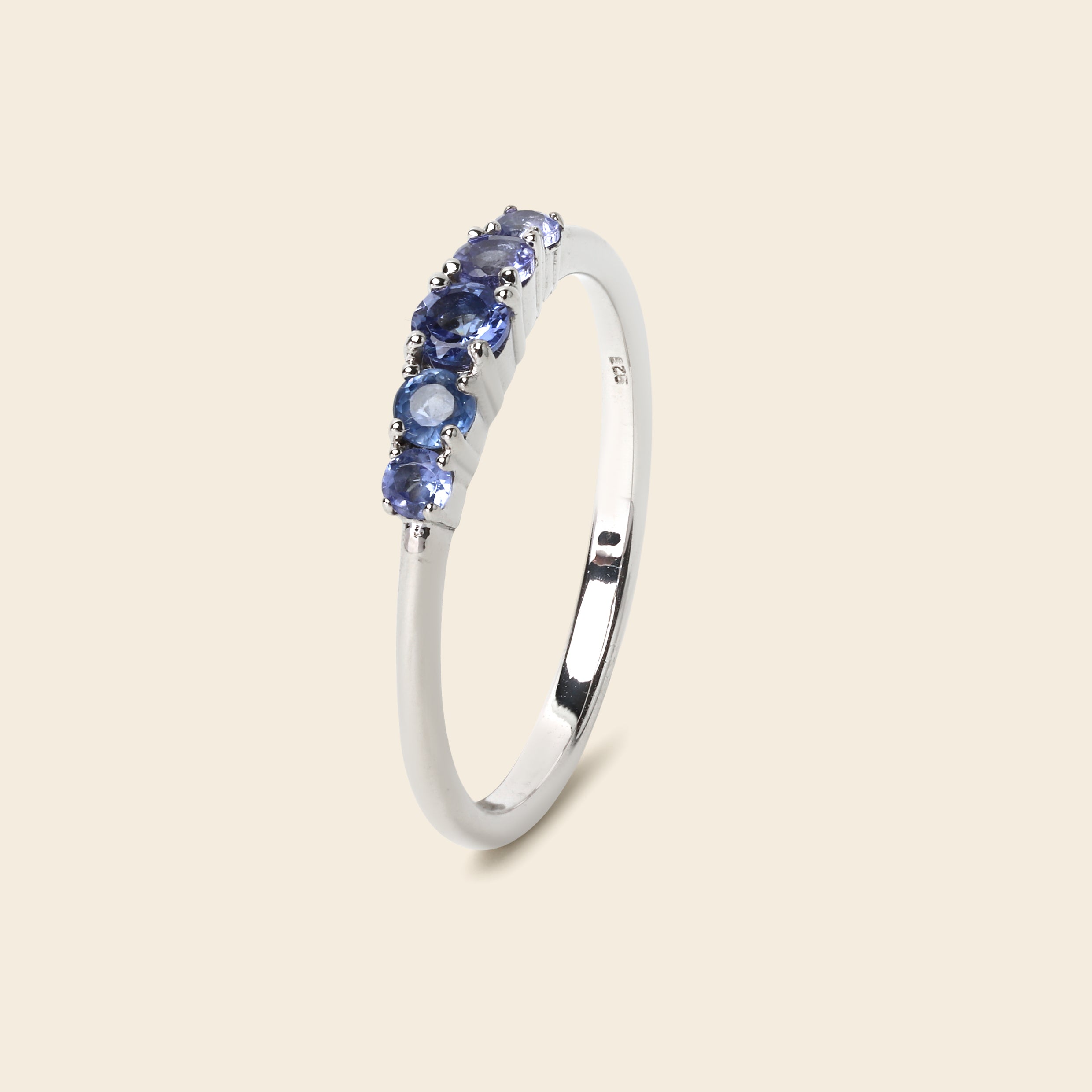 A silver ring featuring a row of round-cut blue gemstones, shown on a solid light beige background.