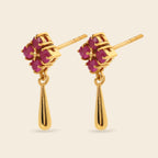 A pair of gold dangle earrings featuring a small cluster of round-cut pinkish-red gemstones in a floral-like setting, with a polished gold teardrop drop hanging below, shown on a light beige background.