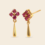 A pair of gold dangle earrings featuring a four-stone floral cluster with deep pinkish-red gemstones and a polished teardrop-shaped drop below, shown on a light beige background.