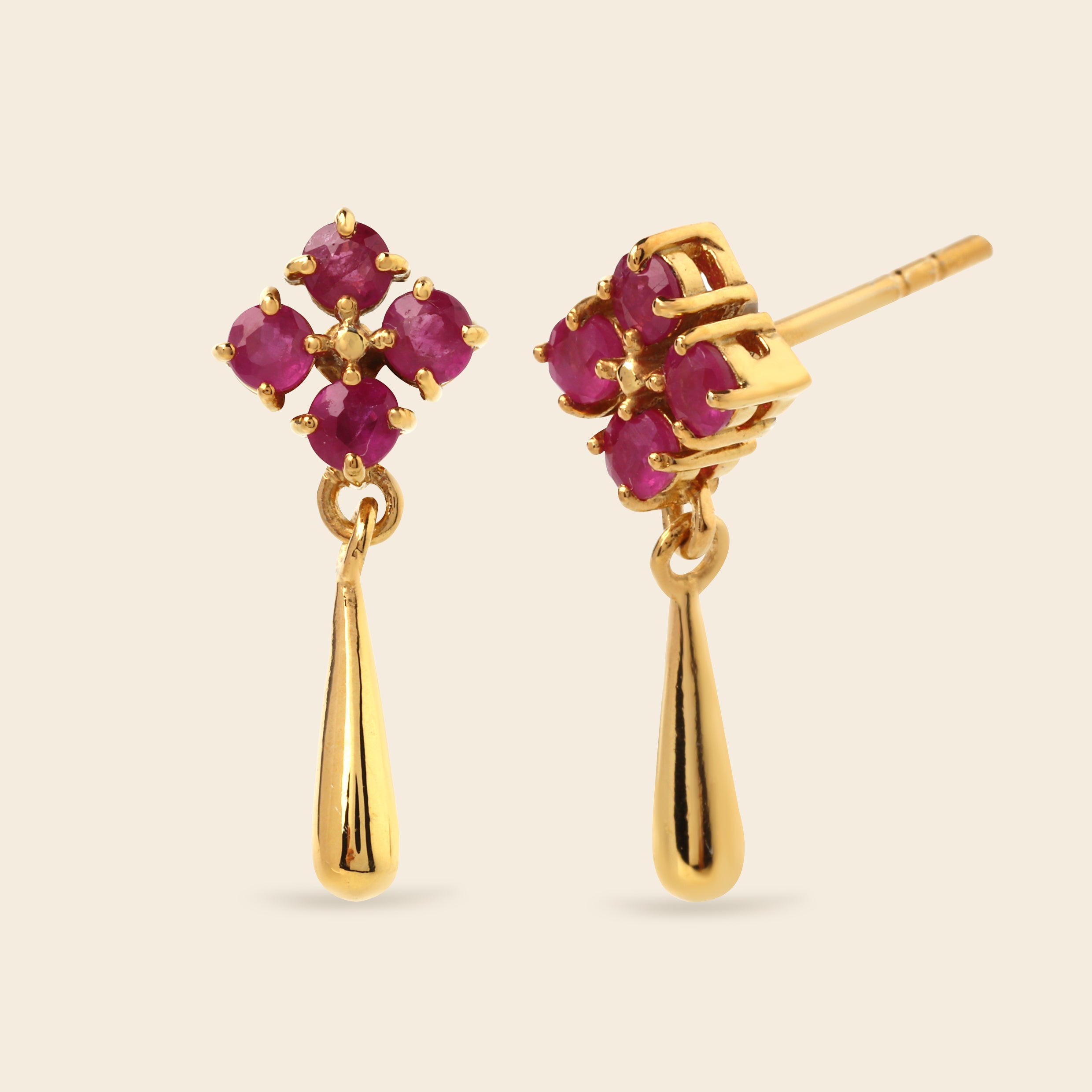 A pair of gold dangle earrings featuring a four-stone floral cluster with deep pinkish-red gemstones and a polished teardrop-shaped drop below, shown on a light beige background.
