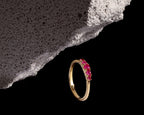 A gold ring with a row of vibrant pink gemstones, displayed on a black background with a textured gray stone prop.