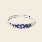 A silver band ring featuring five oval-cut blue gemstones in a row, set against a solid light beige background.