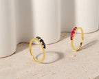 Two gold rings with curved rows of small gemstones, one in dark blue and one in pink, displayed on a light stone surface in front of white cylindrical props.